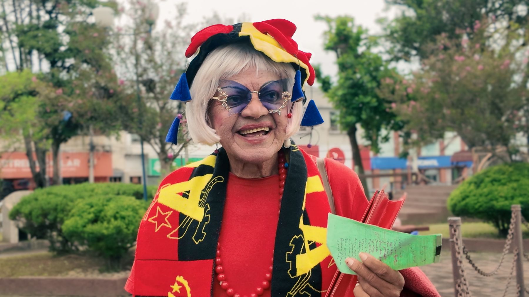 Gui-Gui smiling in a jester hat, blue butterfly glasses and an Angola scarf, in a town square